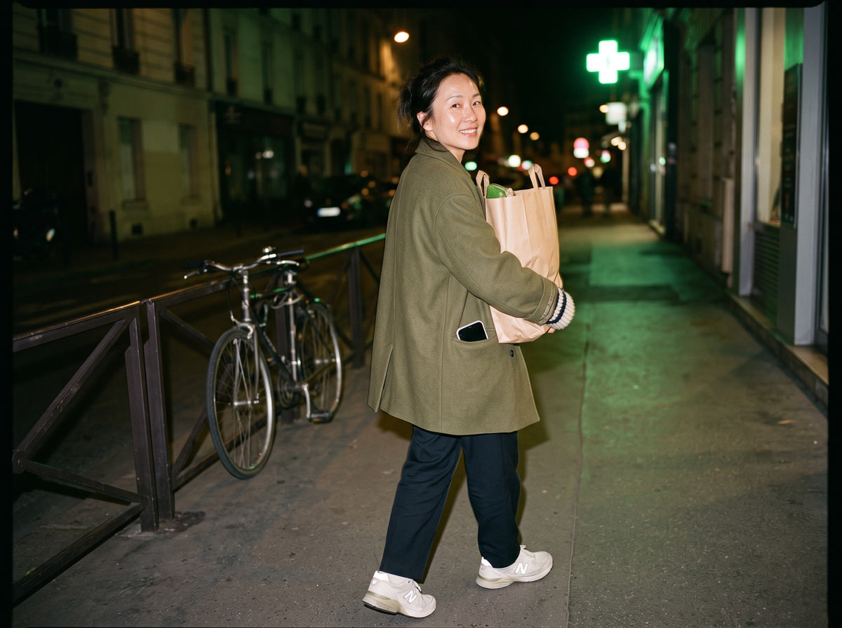 An East Asian woman in her mid 40s, in relaxed tailoring and worn-in trainers, walks home along a city street after dark with a paper bag of groceries held against her hip, looking back over her shoulder and smiling.