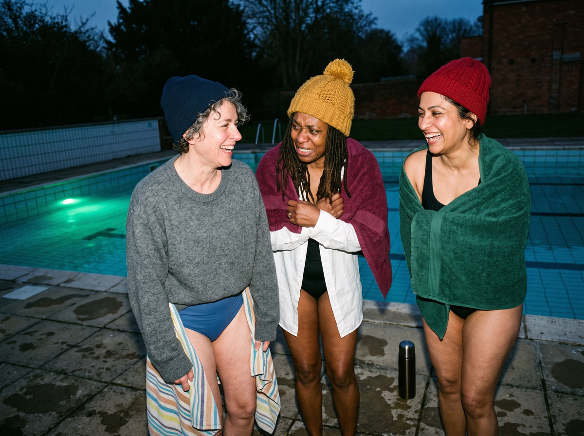 Three women in their late 40s and 50s, of different ethnic backgrounds and different body sizes, in plain swimsuits and woolly hats, stand close together at the edge of an outdoor pool at dusk, wrapped in towels, laughing and shivering.