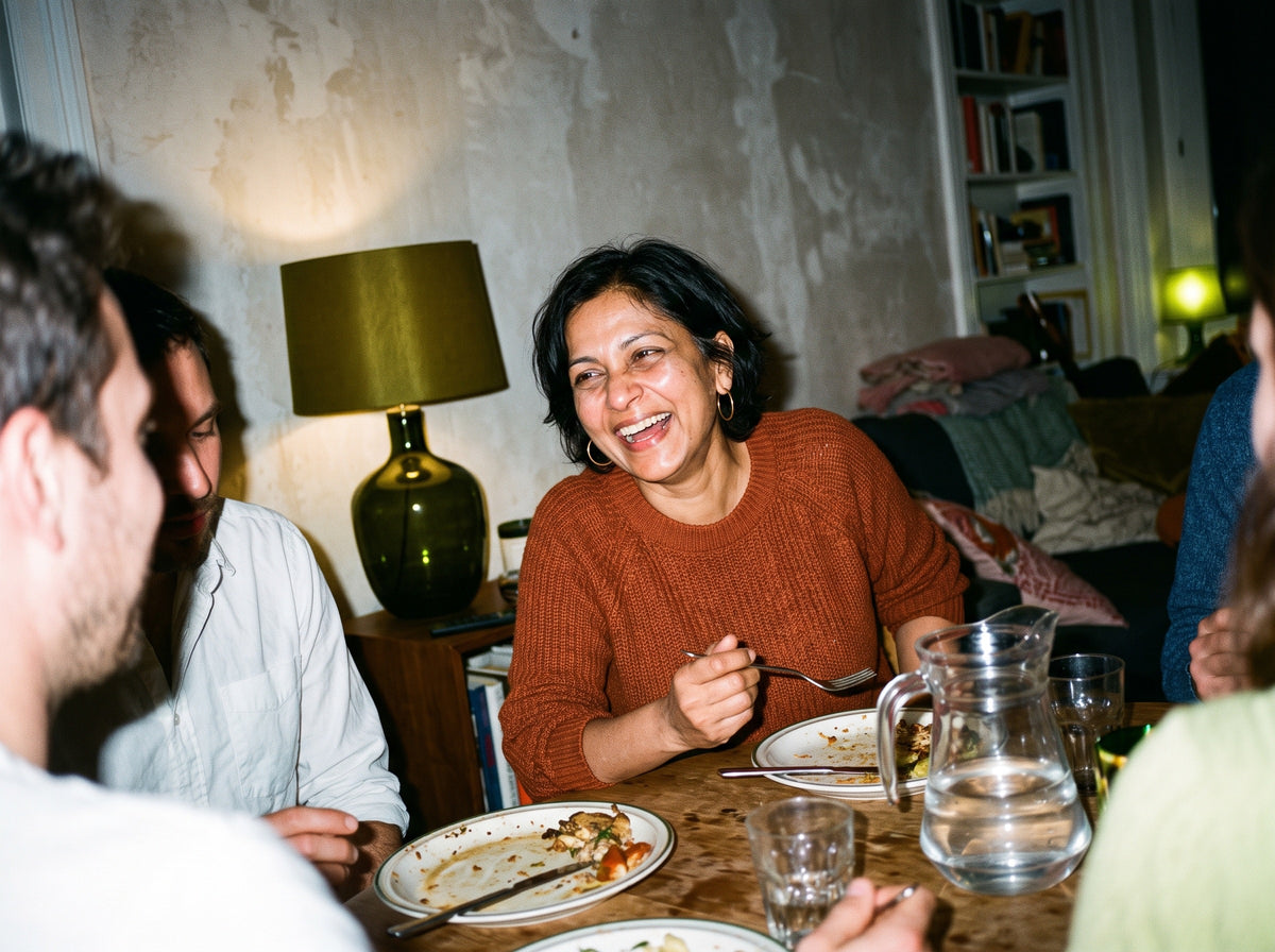 A South Asian woman in her early 50s, in a rust-coloured knit and gold hoop earrings, leans back from a dinner table at a friend's home at night, laughing with her whole face, a fork still in her hand.