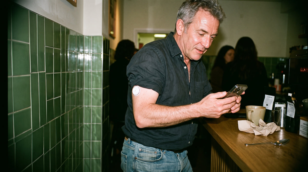 A man in his early 50s waits at a café counter in the morning, glancing at his phone, his rolled sleeve showing a discreet white round glucose sensor on the back of his upper arm.