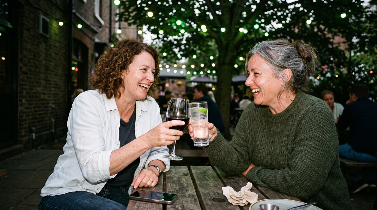 Two women in their 50s at a pub garden table in the evening, clinking a glass of red wine against a tall soda water with lime, mid-laugh.