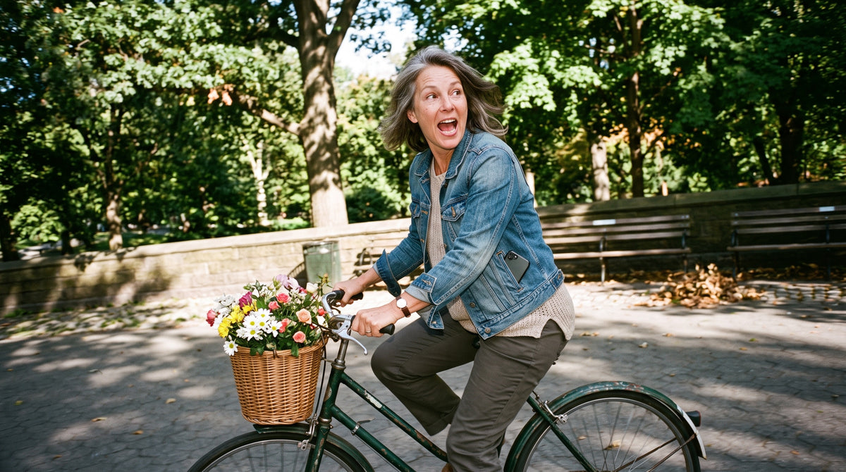 A woman in her mid-50s cycles through a park on an old bicycle with flowers in the basket, turning to shout something to a friend behind her.