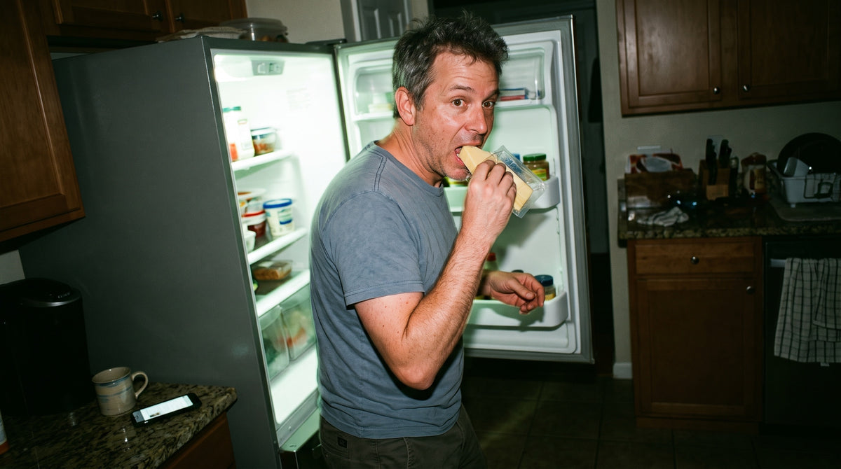 A man in his late 40s stands at an open fridge in a dark kitchen at night in a T-shirt, eating cheese straight from the packet, caught by surprise.