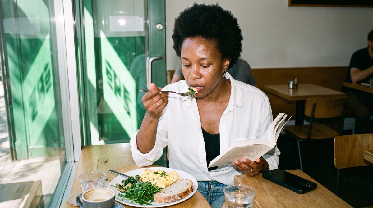 A tall, slim Black woman in her mid 40s with a short natural afro, absorbed and not smiling, eats a proper lunch at a café window seat, eggs, greens and bread on the plate, fork mid-way to her mouth, reading a paperback.